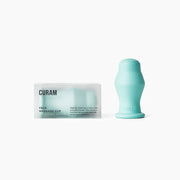 Curam Face Cup i silikon i färgen relaxing green bredvid sin minimalistiska förpackning mot en ren vit bakgrund, som visar ansiktskoppens ergonomiska böjda form och mjuka matta yta.